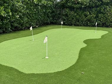 Putting green de grama artificial con tres banderas blancas y un borde de fibra más larga, con un seto detrás.