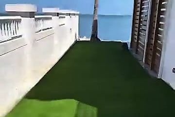 La misma franja con grama artificial verde rematando contra el muro, con el agua detrás.