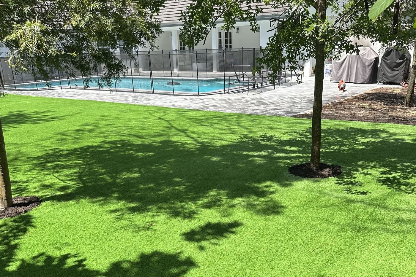 Patio amplio con grama artificial y un árbol en su alcorque, con la piscina vallada y la casa al fondo.