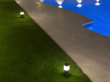 Patio con piscina iluminada de noche y grama artificial junto al pavimento.