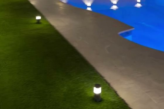 Patio con piscina iluminada de noche y grama artificial junto al pavimento.