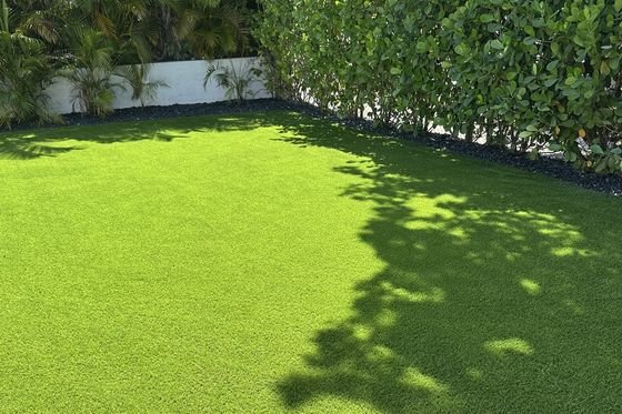 Patio con grama artificial rodeado de un seto alto, con la sombra de un árbol proyectada encima.