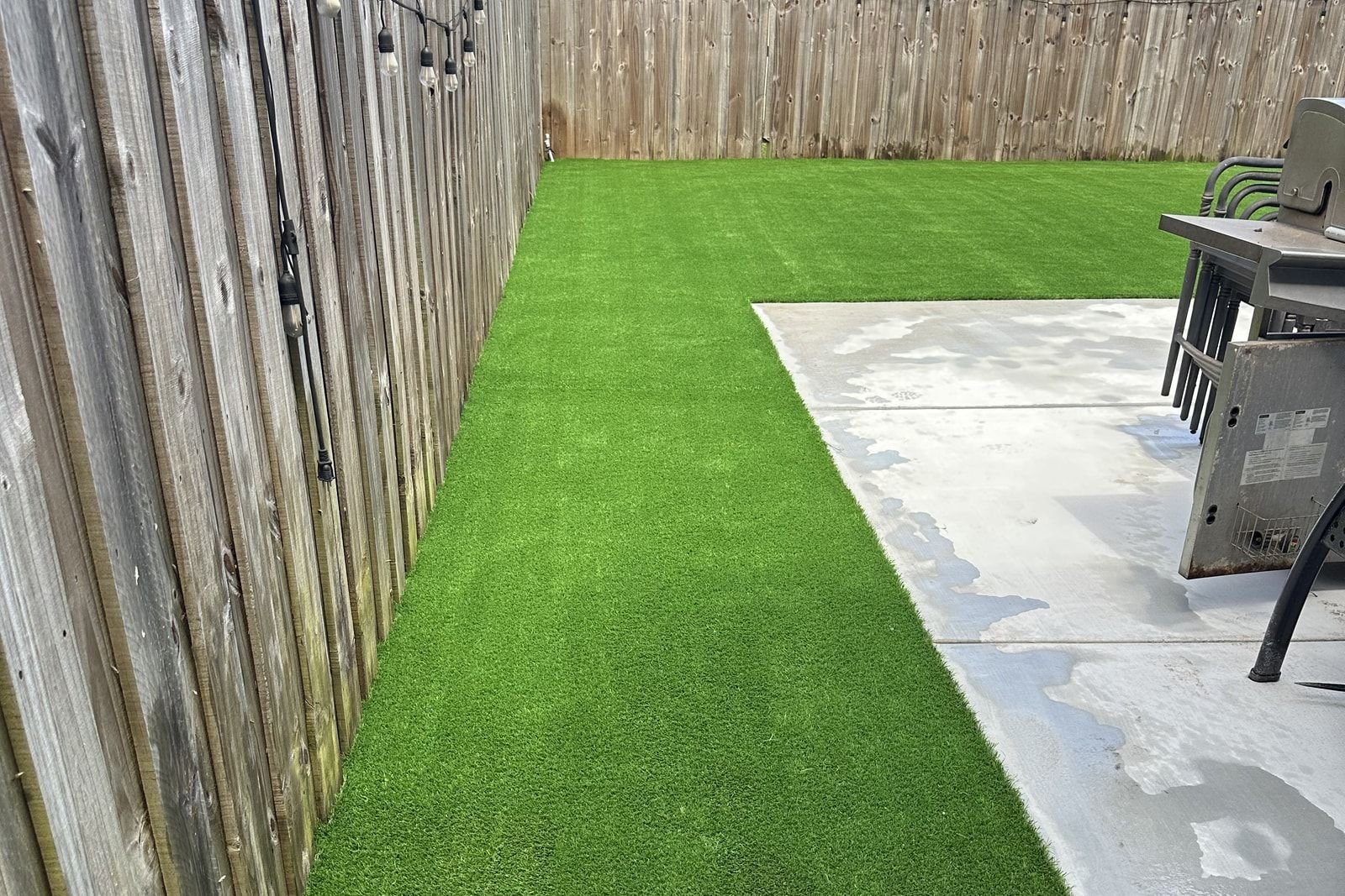 Patio con grama artificial junto a un pavimento de concreto, cerrado por una valla de madera con guirnaldas de luces.