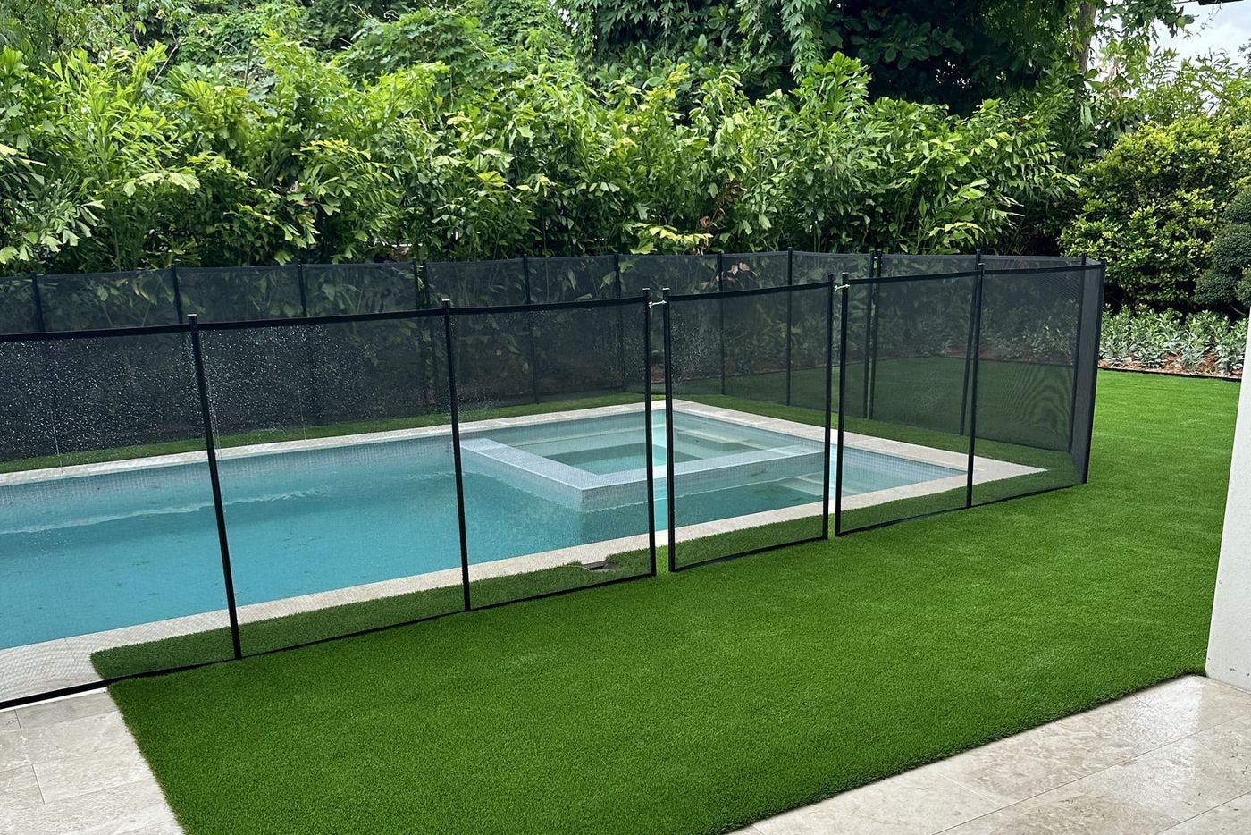 Rectángulo de grama artificial embutido en un pavimento de travertino, junto a una piscina con reja negra.