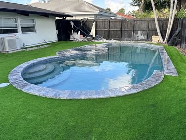Patio con grama artificial rodeando una piscina curva con borde de piedra, junto a una casa de una planta.