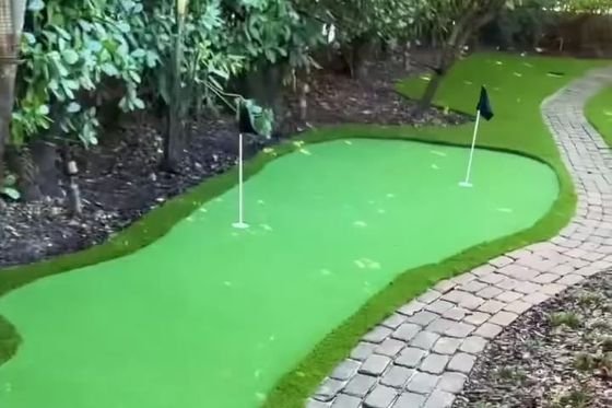 Putting green de grama artificial con dos banderas, junto a un camino curvo de adoquines.